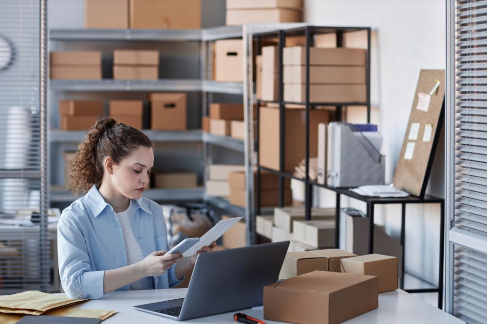 The image displays a focused woman in a light blue shirt, working in a storage room filled with shelves stocked with cardboard boxes. She is examining a document closely while sitting at a desk with a laptop open in front of her, surrounded by open parcels and envelopes. The setting suggests that she is in the midst of processing shipments or handling logistics tasks. This scene is typical of an environment where business mailing solutions are implemented, combining the management of physical goods with digital tracking and coordination to ensure efficient delivery and dispatch of business correspondence and packages. The orderly arrangement of shelves and the presence of mailing supplies underscore the importance of organization in mailing operations for businesses.